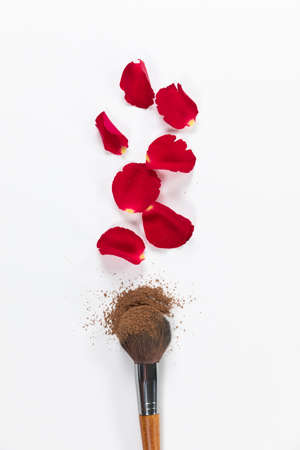 Close up top view of a makeup brush covered with brown powder and red rose petals arranged on a plain white backgroundの写真素材