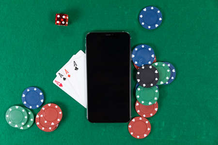 Close up top view of a black smartphone, playing cards, a red dice and colorful gambling chips, arranged on a plain green surface.の写真素材