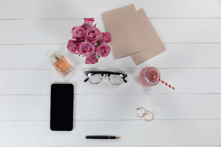 Top view of a smartphone, sheets of paperboard, a pen, earrings, glasses and a smoothie with pink...