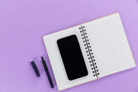 Close up top view of a black smartphone, a notebook and a black pen arranged on a plain purple backgroundの写真素材