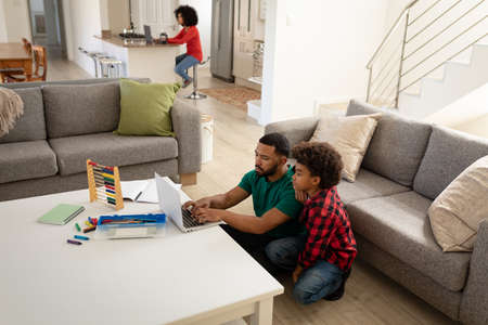 Mixed race couple with son enjoying family time together at home, social distancing and self isolation in quarantine lockdown, father and son are using a laptop and mother is sitting in kitchenの写真素材