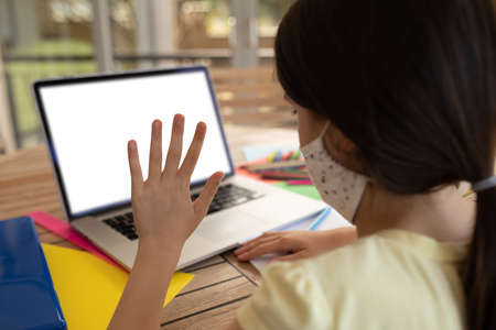 Mixed race girl spending time at home, sitting by table, using laptop, waving. Social distancing
during Covid 19 Coronavirus quarantineの写真素材