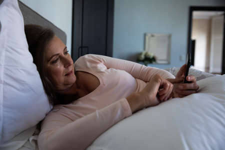 Caucasian woman enjoying time at home, social distancing and self isolation in quarantine lockdown, lying in bed, using a smartphone in bedroom.の写真素材