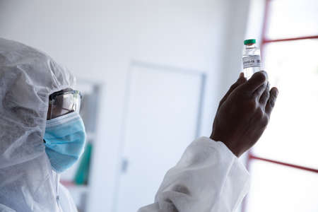 African american male doctor scientist wearing protective clothing and face mask, holding a bottle...