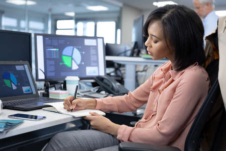 Mixed race businesswoman working in a modern, sitting at desk with computers, writing with pen in...