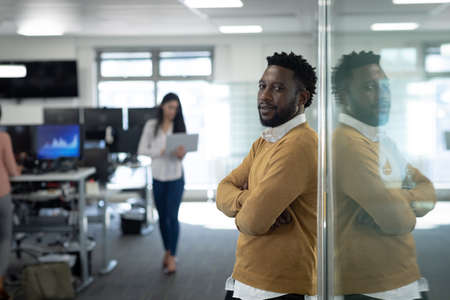 Portrait of African American businessman in a modern office, leaning against glass wall, a colleague in the background. Social distancing in workplace during Coronavirus Covid 19 pandemic.の写真素材
