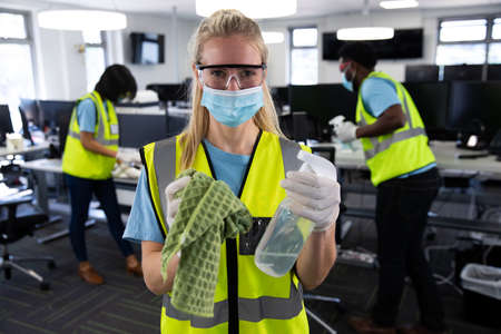 Portrait of Caucasian woman wearing hi vis vest, gloves, safety glasses and face mask, sanitizing office with colleagues in the background. Hygiene in workplace during Coronavirus Covid 19 pandemic.の写真素材