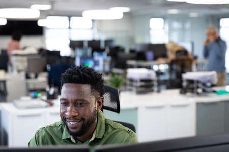 African American businessman working in modern office, sitting at desk looking at computer screen and smiling. Social distancing in workplace during Coronavirus Covid 19 pandemic.の写真素材