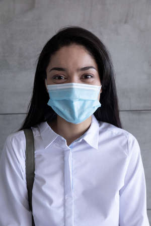 Portrait of mixed race businesswoman wearing face mask working in a modern office, standing in corridor looking to camera. Hygiene, social distancing in workplace during Coronavirus Covid 19 pandemic.の写真素材