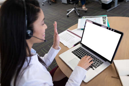 Mixed race businesswoman in modern office, sitting at table using headset and laptop computer for...