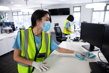Mixed race woman and colleague wearing hi vis vests, gloves and face masks sanitizing an office using disinfectant. Hygiene in workplace during Coronavirus Covid 19 pandemic.の写真素材