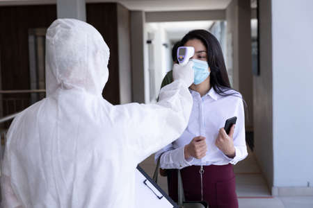 Mixed race businesswoman having temperature taken by person in protective suit before being allowed...