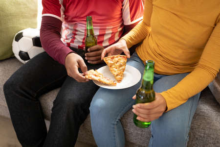 Two African American man at home sitting on sofa in living room, watching sports on TV eating pizza,...