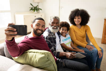 Multi generation African American family at home sitting on sofa in living room, taking selfies with...