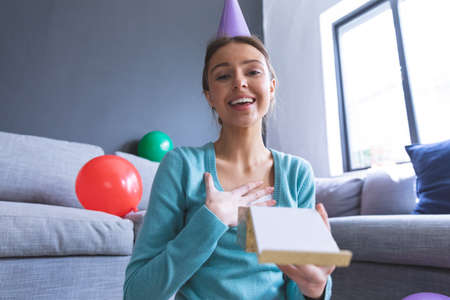 Portrait of Caucasian woman at home celebrating her birthday, in living room wearing party hat, smiling and holding an open gift box. Social distancing during Covid 19 Coronavirus quarantine lockdown.の写真素材