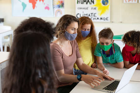 Caucasian female teacher wearing face mask at school, teaching children at school classroom....
