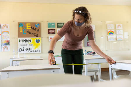 Caucasian female teacher wearing face mask at school, measuring space between desks. Education back...