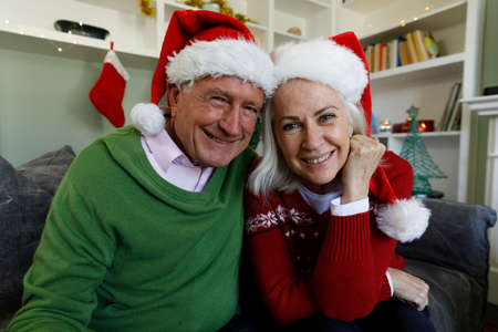 Portrait of senior couple in santa hat smiling while sitting on couch at home. social distancing...