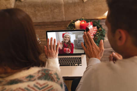 Rear view of couple waving while having a videocall with woman in santa hat waving while holding gift box on laptop at home. social distancing during christmas festivity quarantine lockdown concept.の写真素材