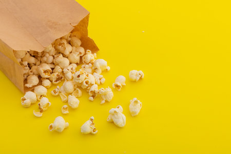 High angle view of popcorn spilling out of brown paper bag on yellow background. fun party food sweet treat snack copy space concept.の写真素材