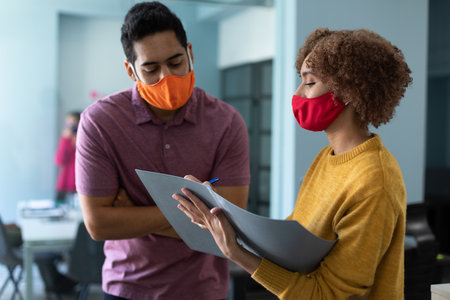 Mixed race man and woman wearing masks in an office. standing, looking at paperwork and talking. hygiene in workplace during coronavirus covid 19 pandemic.の写真素材