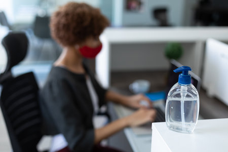 Mixed race woman wearing face mask in an office. sitting and using her computer. hygiene in workplace during coronavirus covid 19 pandemic.の写真素材