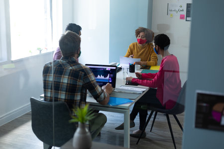 Diverse group of work colleagues wearing masks in an office. sitting and talking together, one man...