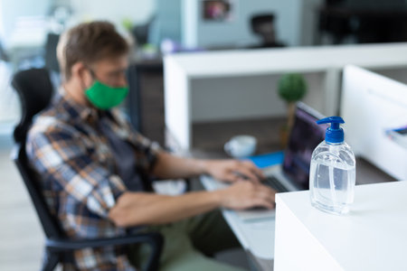Caucasian man wearing face mask in an office. sitting and using his computer. hygiene in workplace during coronavirus covid 19 pandemic.の写真素材