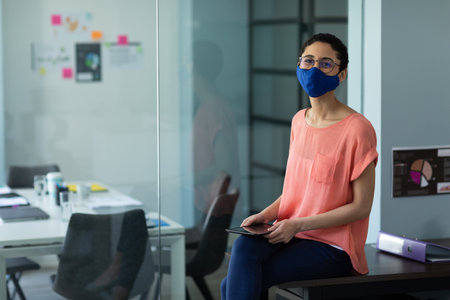 Mixed race woman wearing face mask in an office. sitting, looking at the camera and using digital tablet. hygiene in workplace during coronavirus covid 19 pandemic.の写真素材