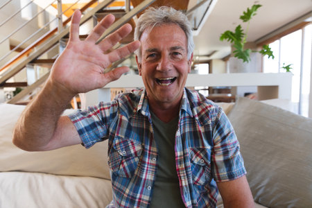 Senior caucasian man laughing and waving during video call. sitting in living room at home. self isolation during coronavirus covid 19 pandemic.の写真素材