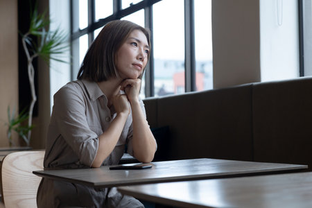 Pensive asian woman looking out of window in office cafeteria. social distancing in business office workplace during covid 19 coronavirus pandemic.の写真素材