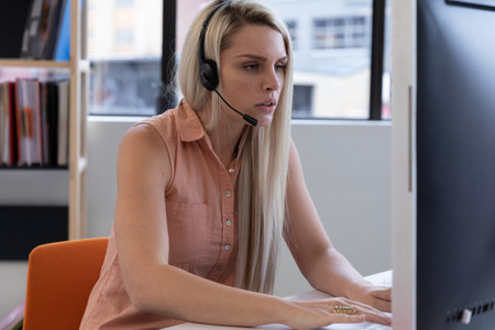 Caucasian woman wearing phone headset using computer in modern office. communication and social distancing in business office workplace during covid 19 coronavirus pandemic.の写真素材