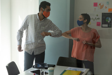 Mixed race man and woman wearing masks bumping elbows in greeting. hygiene in workplace during...