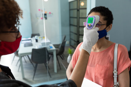 Mixed race woman wearing face mask checking temperature of female colleague. hygiene in workplace...