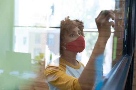 Mixed race woman wearing face mask drawing chart on glass wall in office. hygiene in workplace during coronavirus covid 19 pandemic.の写真素材