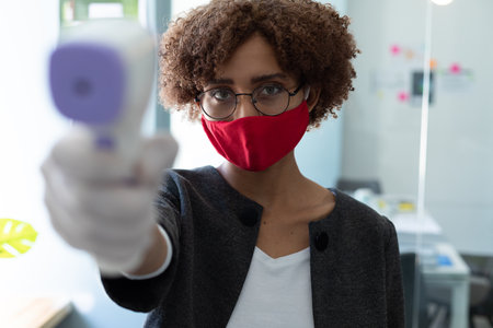 Mixed race woman wearing face mask using thermometer to check health of colleagues. hygiene in workplace during coronavirus covid 19 pandemicの写真素材