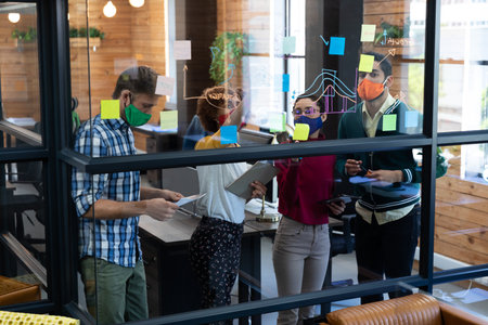 Diverse group of colleagues wearing face masks brainstorming at glass wall in office. hygiene in workplace during coronavirus covid 19 pandemic.の写真素材