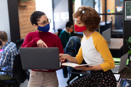 Two mixed race women wearing face masks using laptop and discussing in office. hygiene in workplace...