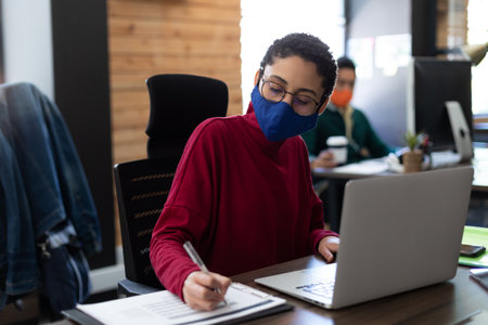 Mixed race woman wearing face mask using laptop and writing in office. hygiene in workplace during coronavirus covid 19 pandemic.の写真素材