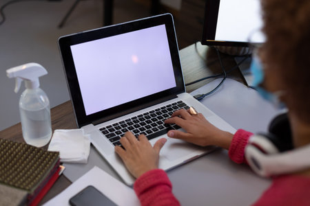 Mixed race woman wearing face mask using laptop at desk in casual office. sanitizing gel on her desk. hygiene in workplace during coronavirus covid 19 pandemic.の写真素材