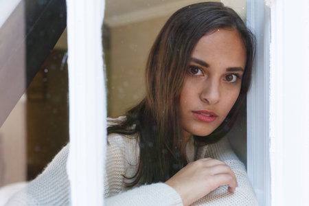 Portrait of caucasian woman looking through window to camera. self isolation at home during covid 19 coronavirus pandemic.の写真素材