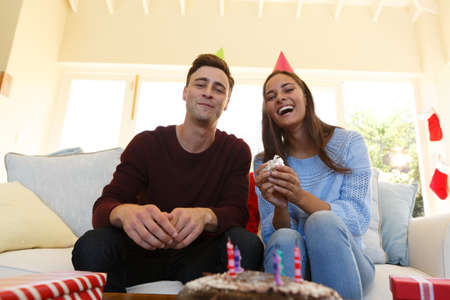 Caucasian couple having birthday video call wearing party hats and holding gift. sitting on couch, birthday cake in foreground.の写真素材