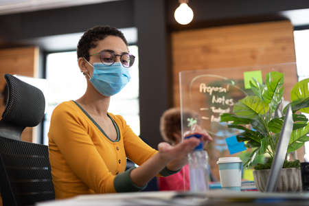 Mixed race woman wearing face mask sanitizing hands at desk in office. colleague in background hygiene screen between them.の写真素材