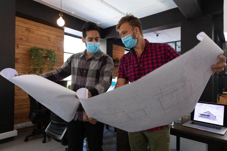 Diverse male colleagues wearing masks in disucssion holding architectural plans in office. hygiene...