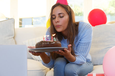 Caucasian woman having birthday video call blowing out candles on cake wearing party hat. self isolation at home during covid 19 coronavirus pandemic.の写真素材