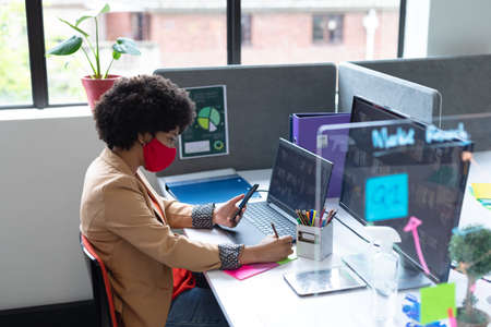 Mixed race businesswoman wearing face mask in creative office. woman sitting at desk, using laptop computer. social distancing protection hygiene in workplace during covid 19 pandemic.の写真素材