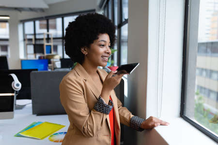 Mixed race businesswoman working in creative office. woman looking through window and talking on smartphone. social distancing in workplace during covid 19 pandemic.の写真素材