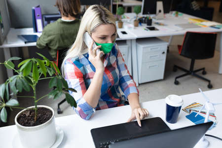Caucasian businesswoman wearing face mask in creative office. woman sitting at desk, using laptop...