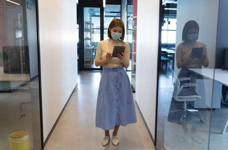 Asian businesswoman wearing face mask standing in corridor using tablet. social distancing health protection hygiene in creative office during covid 19 pandemic.の写真素材