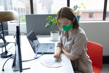 Asian businesswoman wearing face mask sitting at desk looking at samples in creative office. social distancing protection hygiene in workplace during covid 19 pandemic.の写真素材